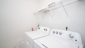 A laundry room with two white front-loading washers and dryers, a metal drying rack on the wall, and a white laundry basket on the floor. - The Rebel Retreat -Oxford's Home Base for Every Rebel Adventure (Oxford)