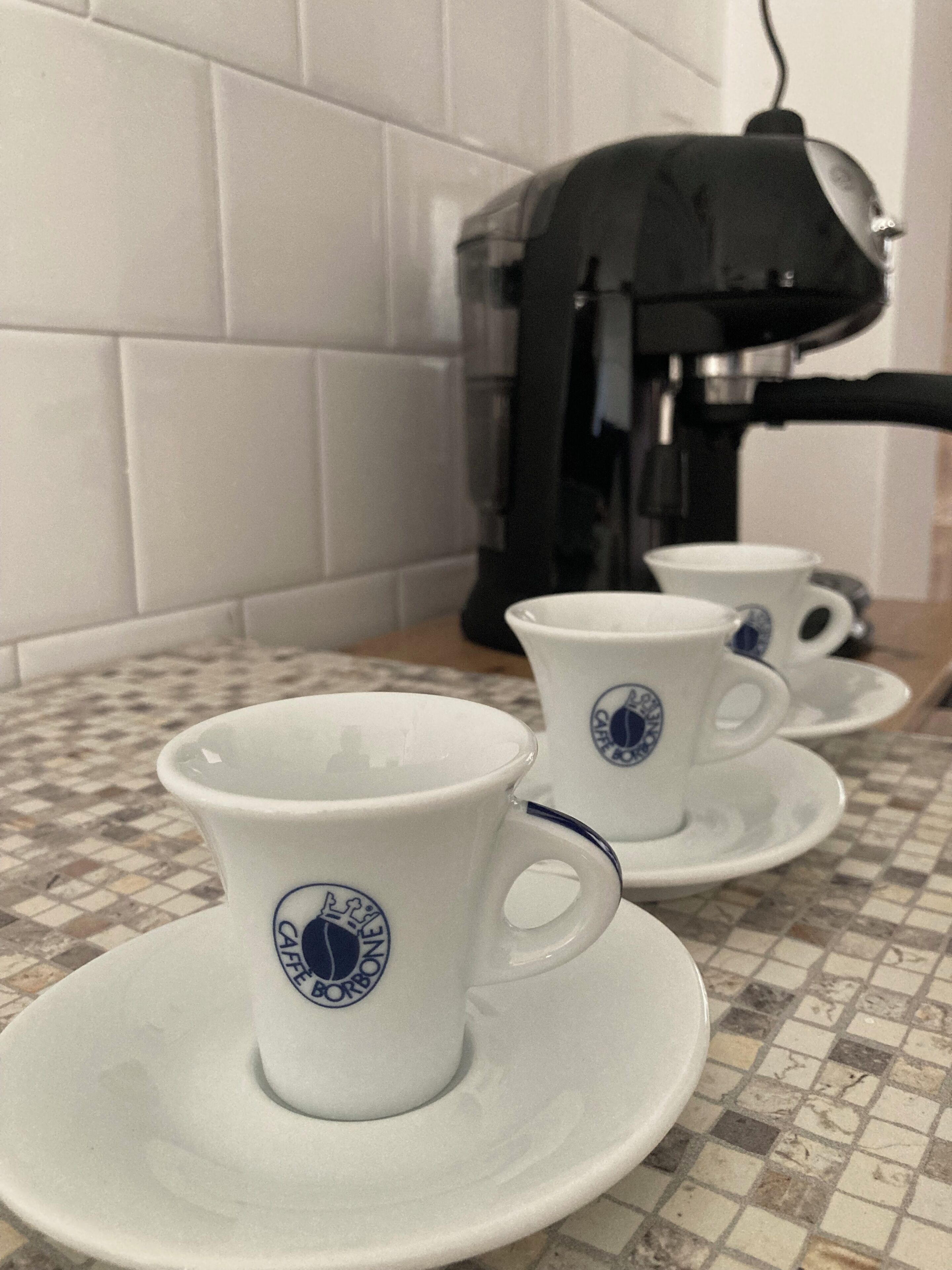 A kitchen area with a coffee maker, two white cups with blue logos, and a tiled backsplash.