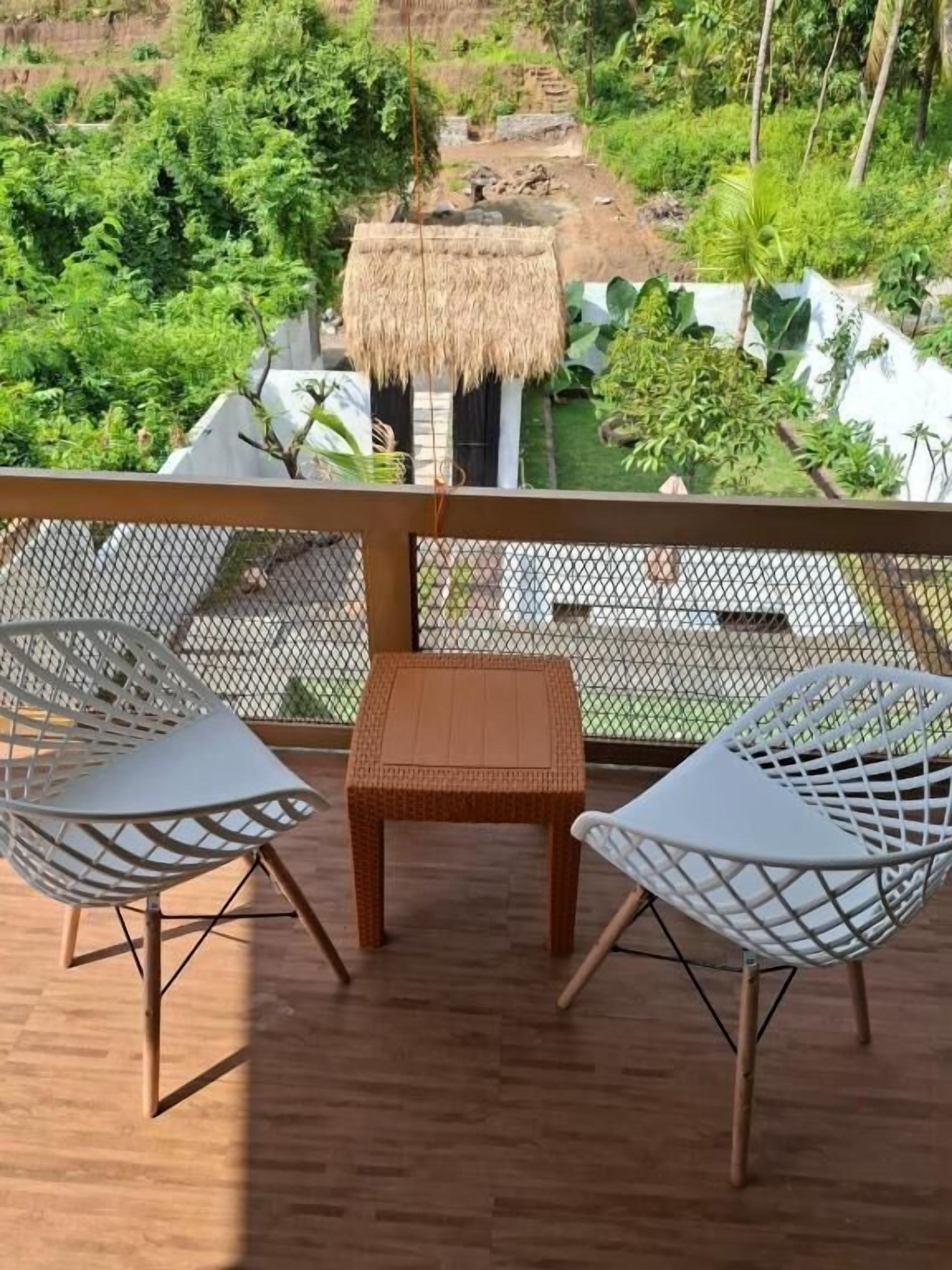 A balcony with wicker chairs and a table, overlooking a lush garden and a thatched-roof structure.
