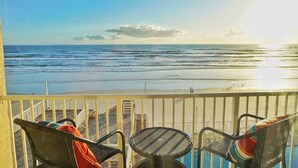Ein Balkon mit zwei Korbstühlen und einem kleinen runden Tisch, der Blick auf einen Strand mit Wellen und einem klaren Himmel bietet.
