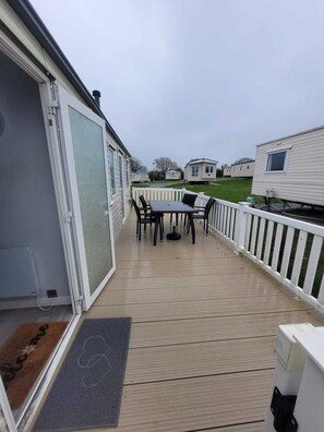 Eine überdachte Terrasse mit Tisch und Stühlen, ein weißer Zaun und im Hintergrund eine Ansicht auf Wohnmobile.