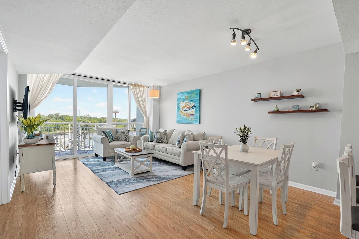 A modern living room with a sofa, dining table, and chairs, a balcony with a view, and a painting on the wall.
