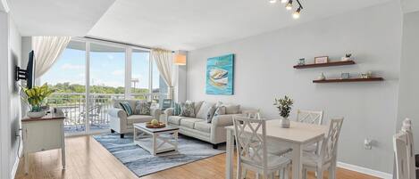 A modern living room with a sofa, dining table, and chairs, a balcony with a view, and a painting on the wall.