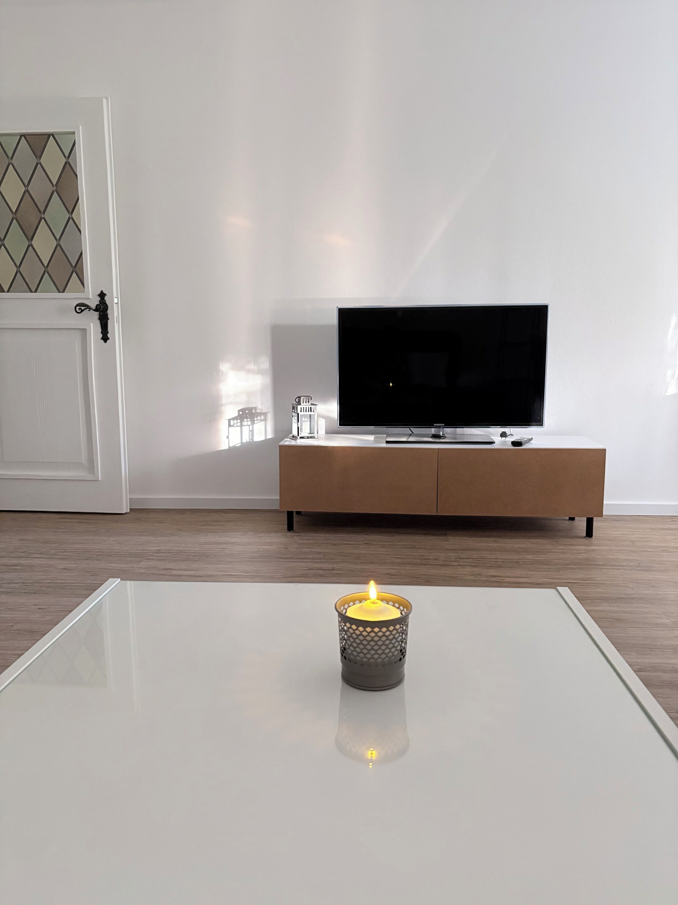 Een moderne woonkamer met een flatscreen televisie op een houten kast, een glazen salontafel met een brandende kaars, en een witte deur met een ruitverdeling in diamantvorm.