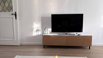 Een moderne woonkamer met een flatscreen televisie op een houten kast, een glazen salontafel met een brandende kaars, en een witte deur met een ruitverdeling in diamantvorm.