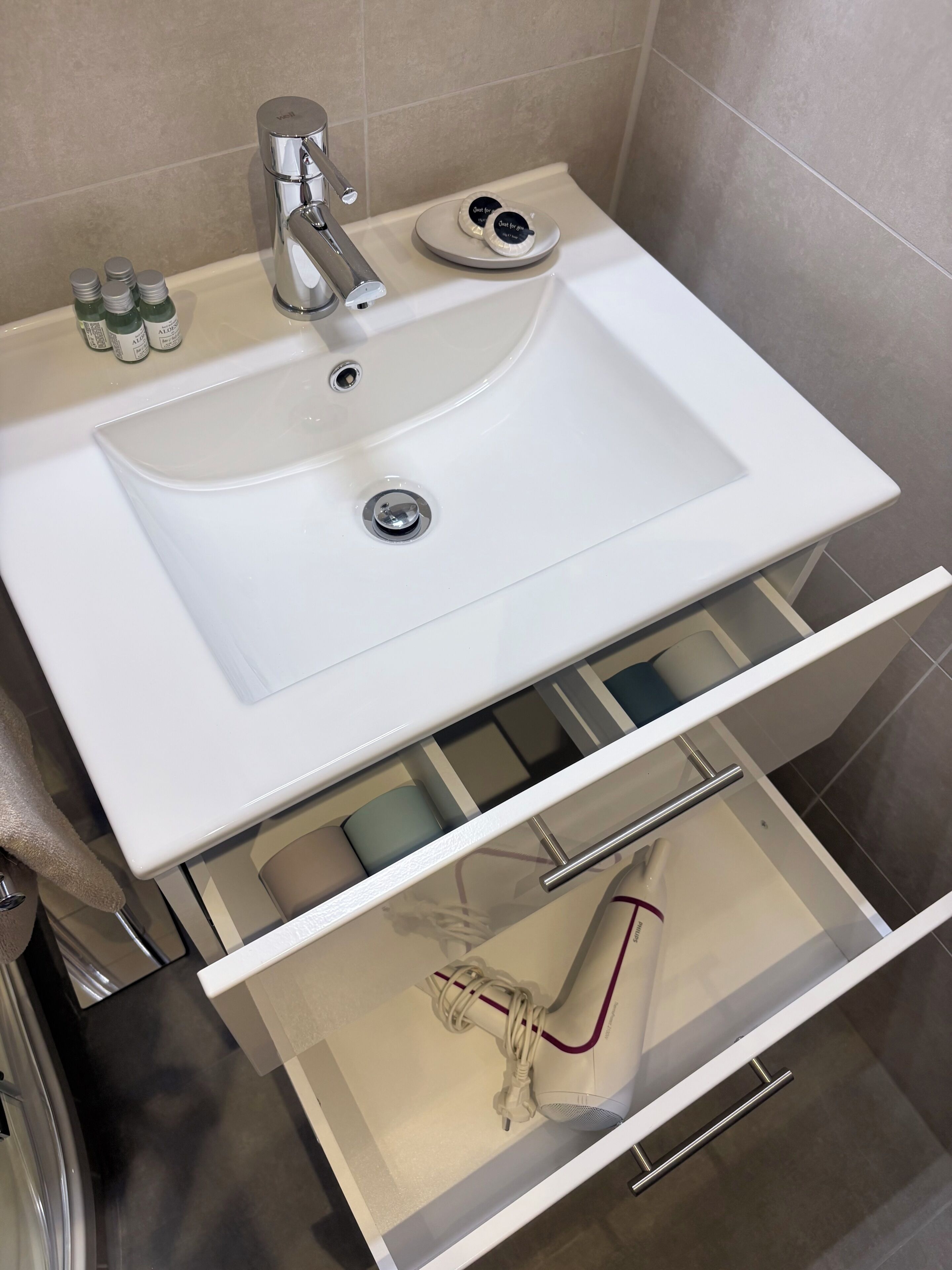 Un baño con lavamanos blanco, grifo de cromo y un cajón con secador de pelo y otros artículos de aseo.
