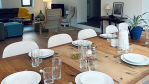 Un comedor con una mesa de madera dispuesta para cuatro personas, rodeada de sillas. Se observa un sofá y una silla al fondo, y una lámpara sobre la mesa.