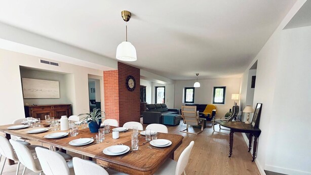 Un comedor con una mesa de madera dispuesta para seis comensales, sillas blancas, una pared de ladrillo decorativa y un reloj.