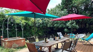 Una terraza de madera con una mesa y sillas, rodeada de follaje y sombrillas rojas.