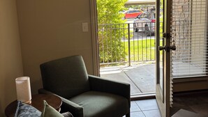 Un soggiorno con un divano, un tavolo e una sedia. Una porta vetrata scorrevole dà accesso a un balcone con vista su auto parcheggiate e aree verdi.