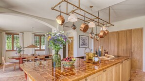 A kitchen with a large island, copper pots hanging from the ceiling, a vase with flowers, and a dining area with a table and chairs. - Villa Sarrazine: Sea View & Walk to Beach (La Croix-Valmer)