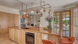 A kitchen with a large island, wooden cabinets, a built-in oven, hanging copper pots, and a view to the outdoors. - Villa Sarrazine: Sea View & Walk to Beach (La Croix-Valmer)