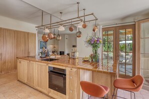 Eine Küche mit einer großen Kochinsel, Holzschränken, einem eingebauten Backofen, hängenden Kupferkesseln und Blick ins Freie.