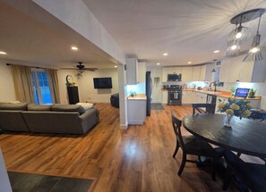 A modern living room with a sofa, a dining table with chairs, a kitchen area with appliances, and a TV mounted on the wall. - Cozy 2-bedroom house in fantastic Oswego with AC and fitness room (OSWEGO)