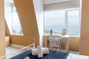 A bedroom with a bed, a desk with a chair, a window with a view, and a bedside table with towels. - Highfield Terrace - Imposing, Beautiful Victorian House With Stunning Sea Views (Ilfracombe)