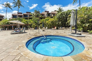A swimming pool with lounge chairs and umbrellas, surrounded by palm trees and a building with balconies. - Fully Remodeled, Lanai Ocean Views, 4.95 SuperHost, 1 br, 2 bath, 3 beds (Kihei)