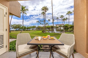 A patio with wicker chairs and a table set with fruit and a pitcher, overlooking a landscaped garden with palm trees and a fountain. - Fully Remodeled, Lanai Ocean Views, 4.95 SuperHost, 1 br, 2 bath, 3 beds (Kihei)