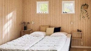 A double bed with a white duvet, two bedside tables with lamps, a small shelf with decorative items, and two windows with plants. - Secluded holiday oasis near lovely beach (Højby)