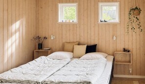 Un lit double avec une couette blanche, deux tables de chevet avec des lampes, une petite étagère avec des objets décoratifs et deux fenêtres ornées de plantes.