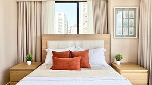 A hotel room with a large bed, a window with curtains, a headboard, and two bedside tables with plants. - City Center| Pool and Gym 1BR by YellowKey (Santo Domingo)