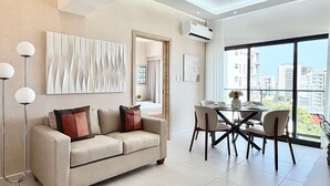 A modern living room with a sofa, dining table, and chairs, a large window with a city view, and an air conditioning unit. - City Center| Pool and Gym 1BR by YellowKey (Santo Domingo)