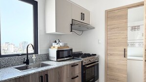 A modern kitchen with a microwave, stove, and sink, wooden cabinets, and a window with a city view. - City Center| Pool and Gym 1BR by YellowKey (Santo Domingo)