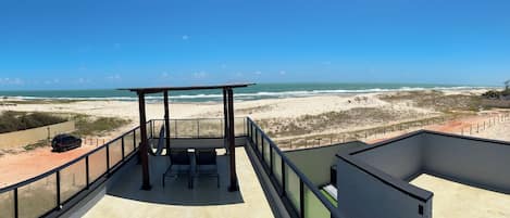A balcony with a view of the beach, a table, and chairs.