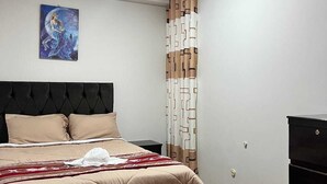 Une chambre à coucher comprenant un lit, une tête de lit, une œuvre d’art accrochée au mur, un rideau à motifs et un plancher en bois.