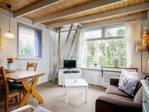 Ein gemütliches Zimmer mit Holzboden, einem Fernseher auf einem weißen Schrank, einem Esstisch mit Stühlen, einem Sofa und einem Fenster mit Vorhängen.