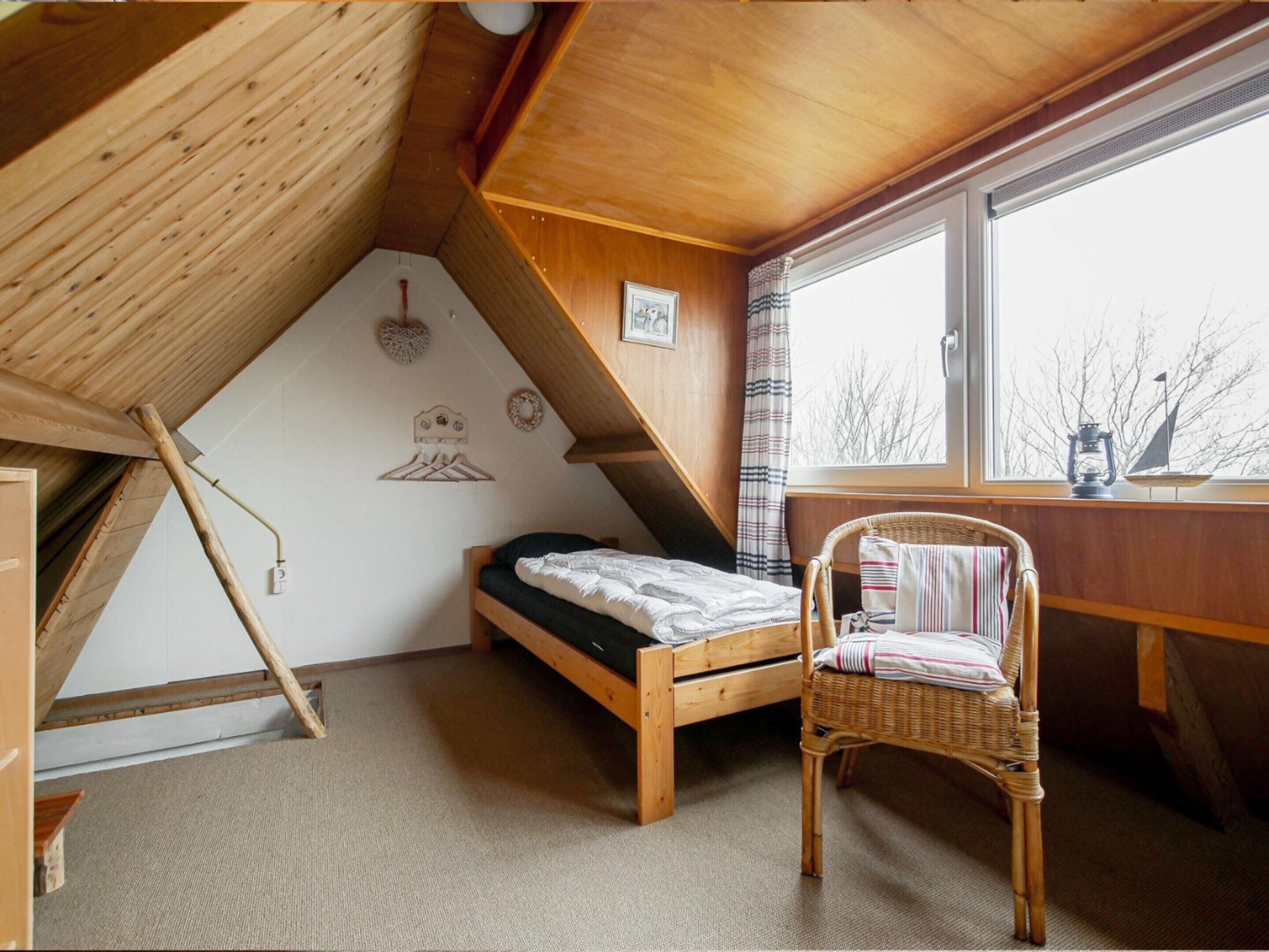 A small, cozy room with a sloped ceiling, a single bed, a wicker chair, and a window with a view of trees.
