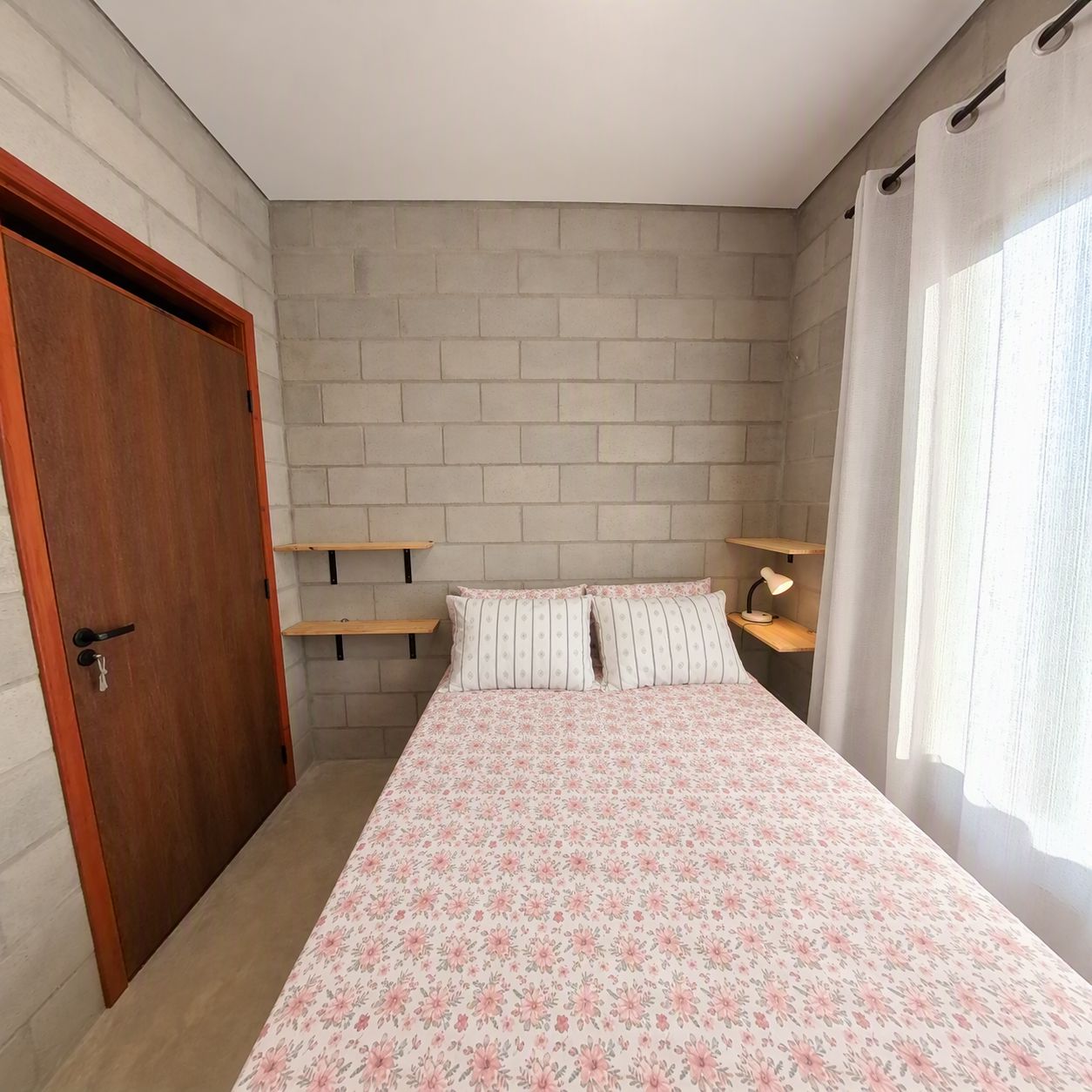 A bedroom with a brick-patterned wall, a wooden door, a bed with floral bedding, and a bedside table with a lamp.