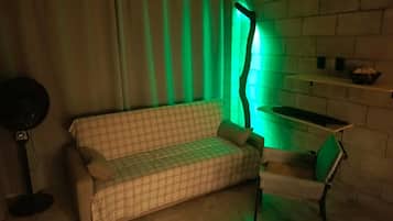 Ein Zimmer mit einer Couch, einem Sessel, einem Ventilator und einem Wandregal.