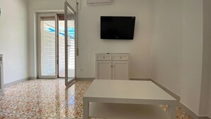 Une pièce dotée d’un sol carrelé, d’une table blanche, d’un téléviseur fixé au mur et d’une porte-fenêtre coulissante donnant sur un espace extérieur.