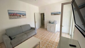 Un salon avec un canapé gris, une table basse blanche, des carreaux de sol à motifs, un coin cuisine avec des placards et un téléviseur fixé au mur.