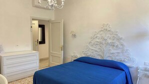 Une chambre à coucher avec un lit, une commode, un lustre et une porte donnant sur une autre pièce.