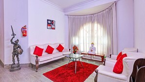 Una sala de estar con un sofá blanco, cojines rojos, una mesa de centro de vidrio, un tapete rojo, una estatua de Buda y un extintor en la pared.