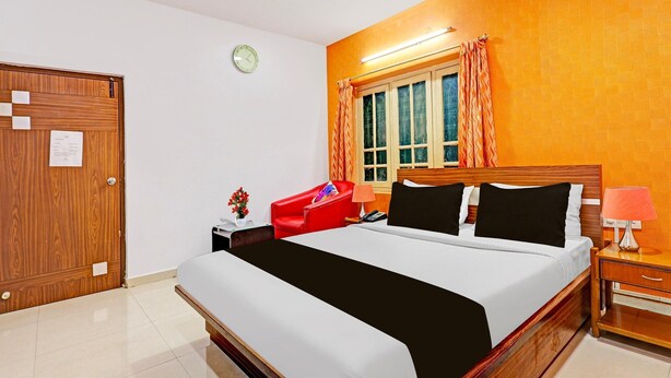 Habitación de hotel con cama, una silla roja, mesita de noche, lámpara, reloj y ventana con cortinas.