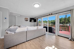 A modern living room with a grey sofa, a mounted TV, and a view of a lake through large sliding doors. - Luxury Lakefront Retreat with Private Dock ,Sleeps 16 (Lake Harmony)