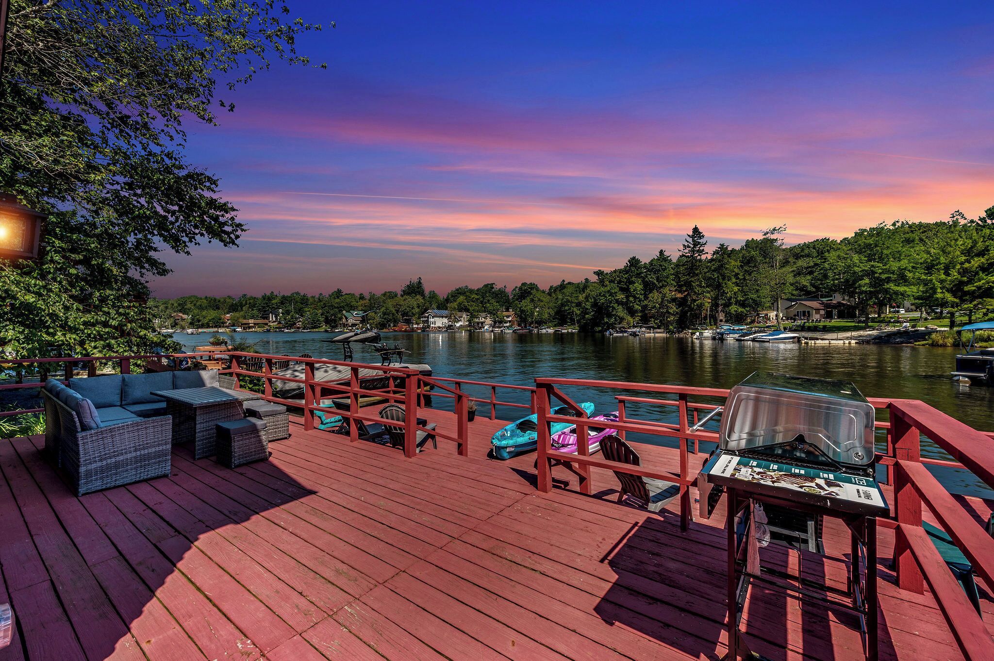 A wooden deck with outdoor furniture, a grill, and boats on a calm lake with a sunset view.