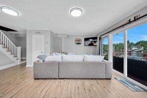 A modern living room with a grey sofa, wooden flooring, a staircase, a door, a wall clock, and a TV. - Luxury Lakefront Retreat with Private Dock ,Sleeps 16 (Lake Harmony)