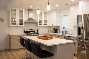 A modern kitchen with white cabinetry, a central island with bar stools, stainless steel appliances, and pendant lighting. - Villas House - 10 mins to Cape May, 15 to Wildwood (Lower Township)