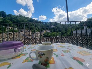 A table with a fruit-patterned tablecloth, a purple lid, and a floral mug, overlooking a village with terracotta roofs and a castle in the distance.