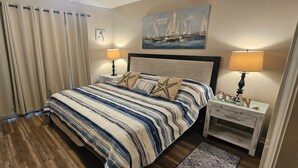Een slaapkamer met een gestreept beddegoed, twee bedlampen, een schilderij van zeilboten en een nachtkastje met een lade.