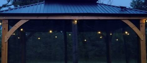 A covered outdoor seating area with a roof, surrounded by a wooden structure, and string lights.