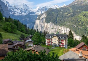 Uma aldeia de montanha com edifícios tradicionais, um hotel em destaque com detalhes vermelhos e picos nevados ao fundo.