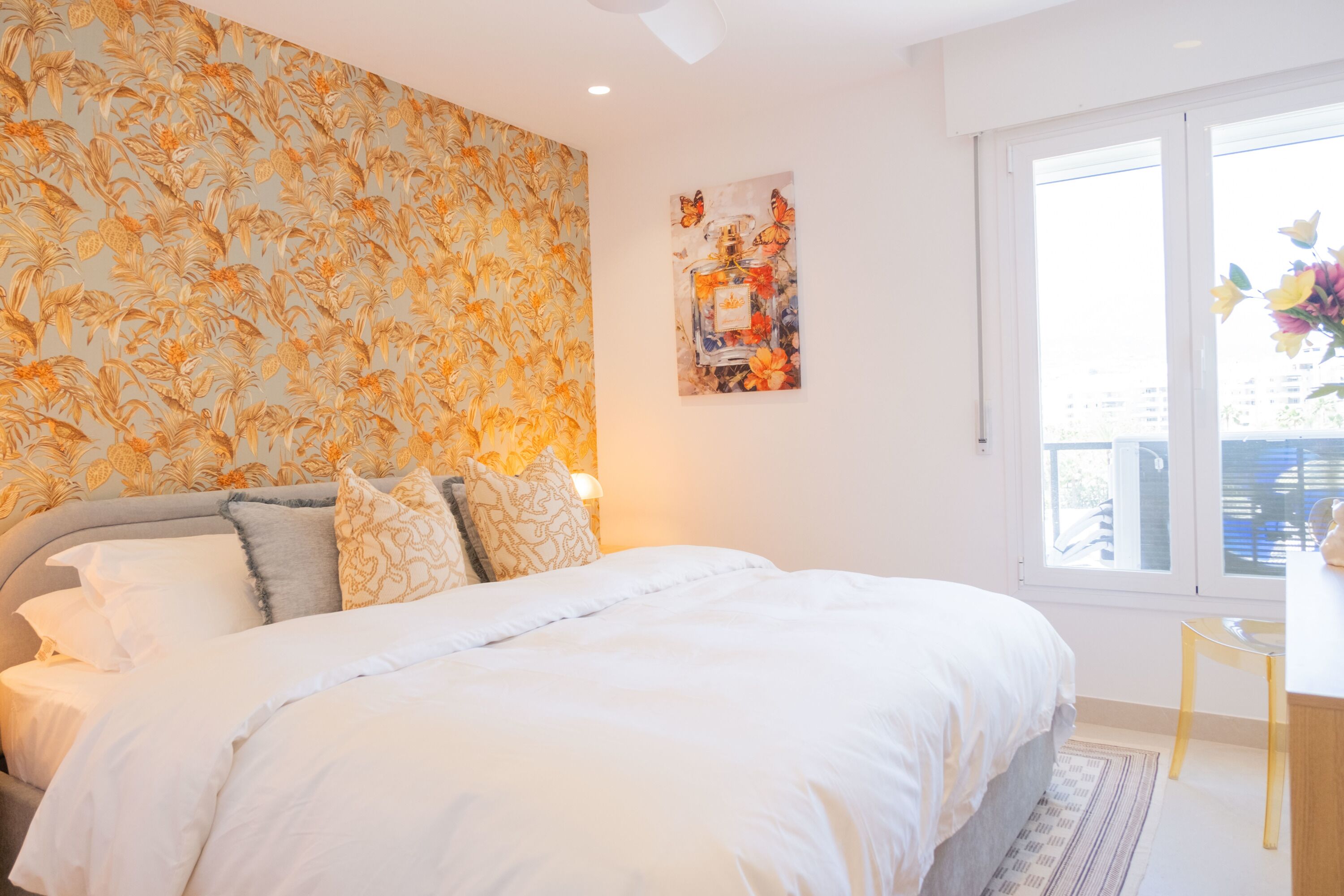 A bedroom with a large bed, a patterned wallpaper, a painting, and a window with a view.