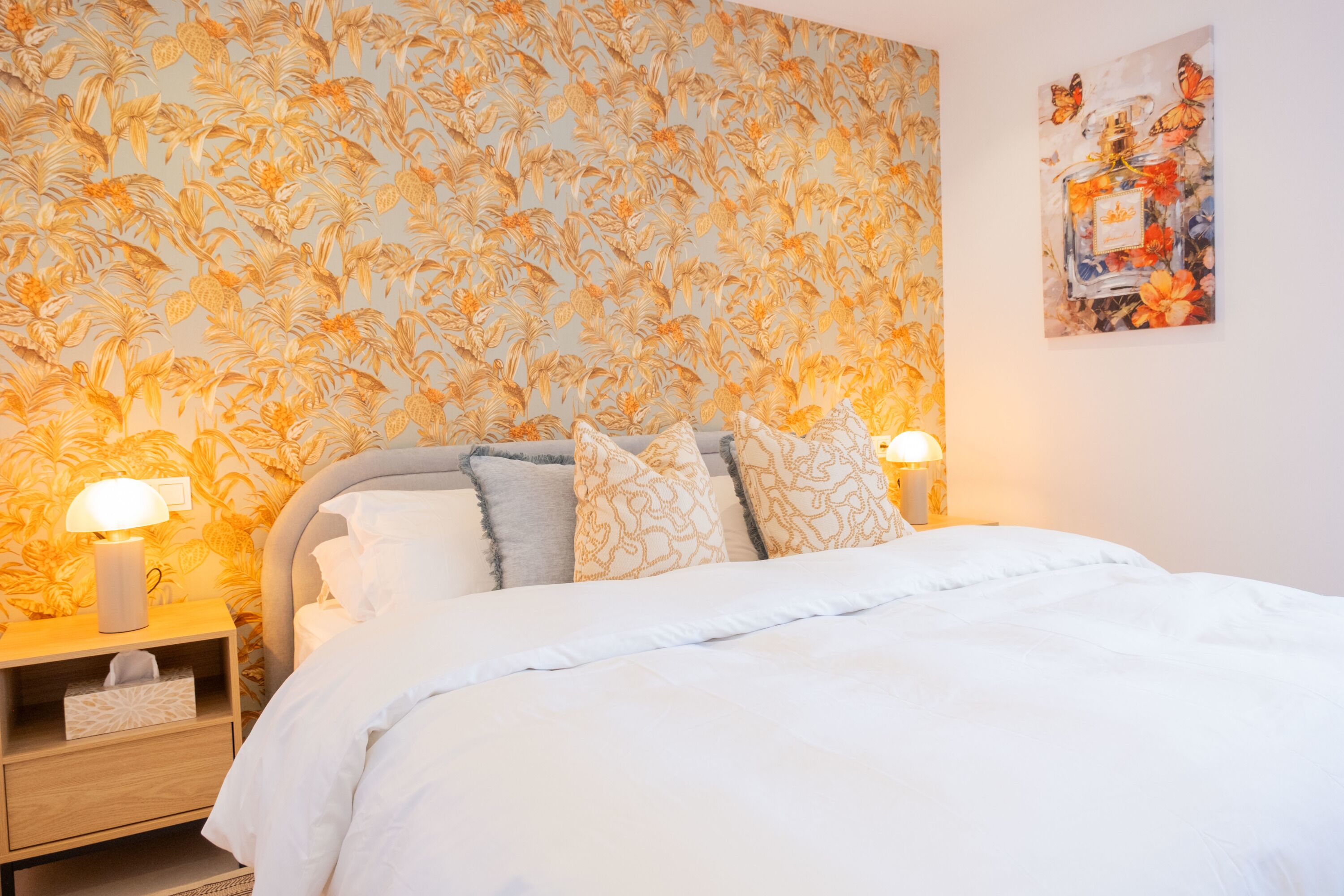 A bedroom with a floral wallpaper, a bed with white bedding, two bedside lamps, and a wall-mounted painting.