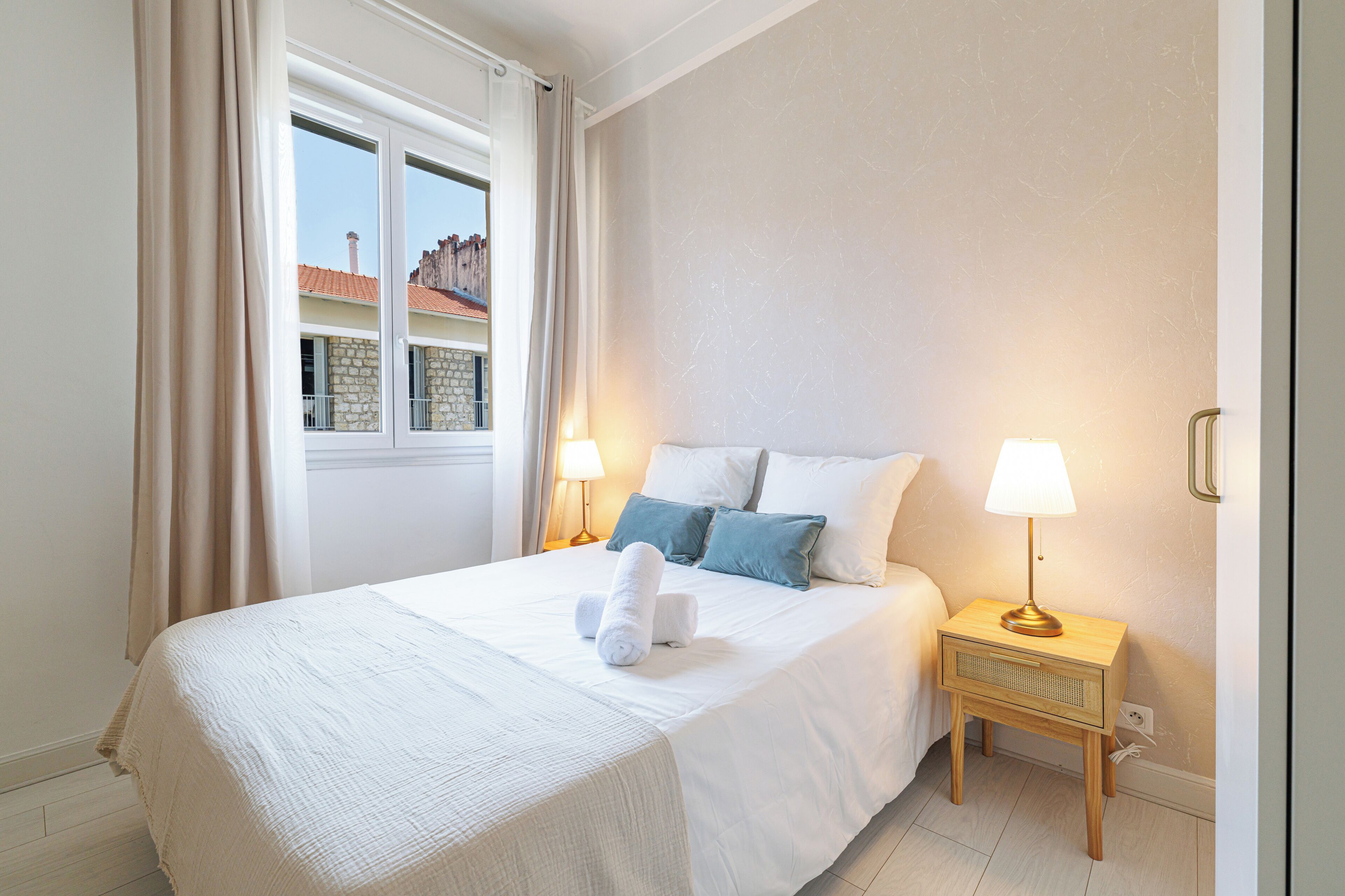 A hotel room with a bed, two bedside lamps, a window with curtains, and a view of buildings outside.