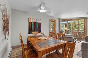 Un coin repas avec une table en bois, des chaises, un ventilateur de plafond et une vue sur un espace extérieur, accessible par une porte vitrée, où se trouvent une chaise et une lampe.