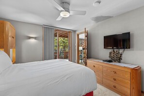 Un dormitorio con cama, ventilador de techo, televisión, cómoda y puerta corrediza a un balcón.